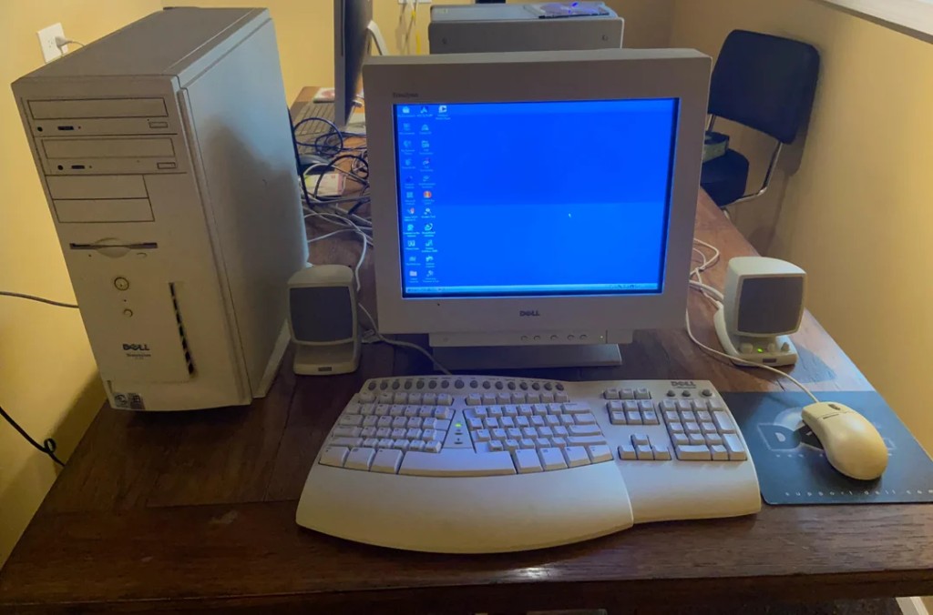 Beige Dell tower, CRT monitor showing a classic Windows desktop, split ergonomic keyboard, mouse, and two small speakers on a wooden desk — the family PC before DSL.